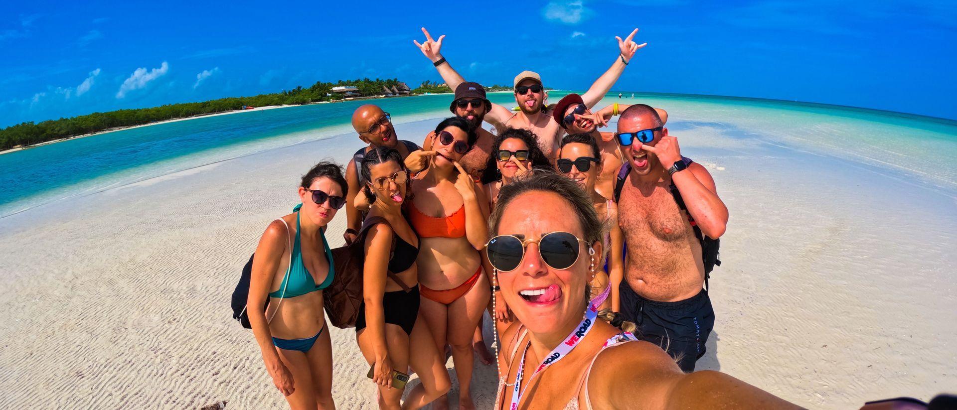 Un gruppo WeRoad si scatta un selfie giocoso su un banco di sabbia bianca, circondato da acque turchesi sotto un cielo azzurro.