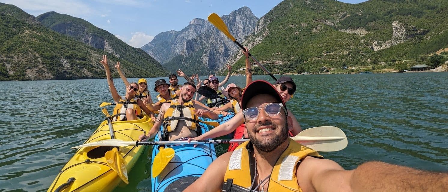 Un gruppo WeRoad sorride per un selfie in kayak su un lago tra imponenti montagne a picco.
