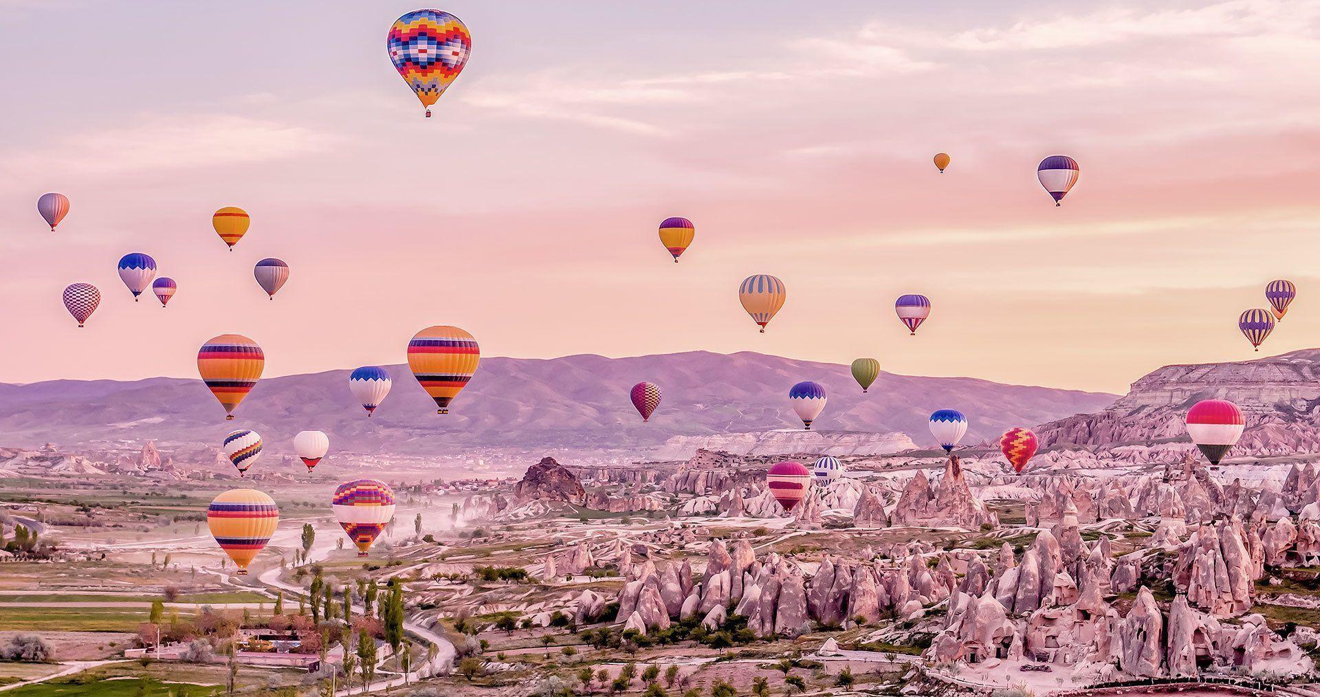De nombreux ballons à air chaud colorés flottent au-dessus d'un paysage de vallée rocheuse avec des montagnes lointaines sous un ciel rose.