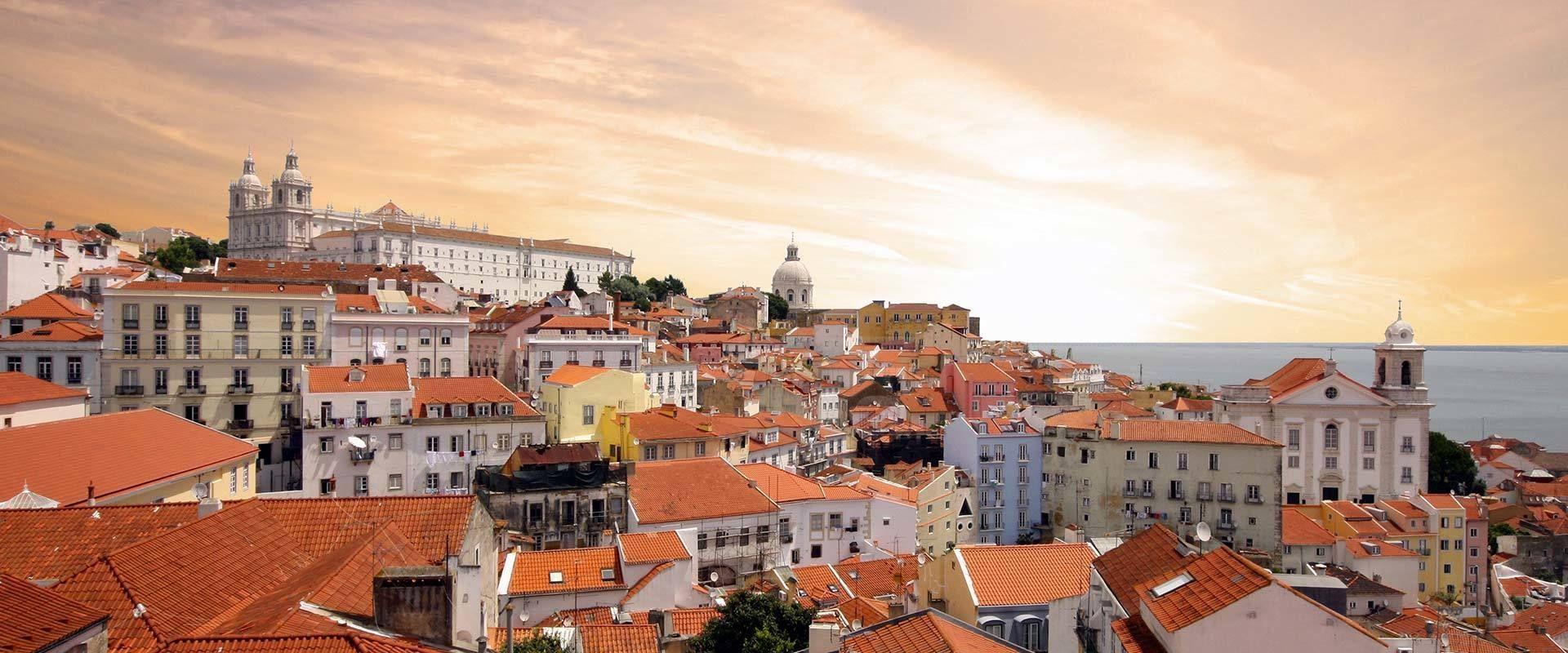 Un paesaggio urbano con tetti di tegole rosse ed edifici colorati che si ergono su una collina, verso un grande monastero, sotto un cielo caldo e nuvoloso.