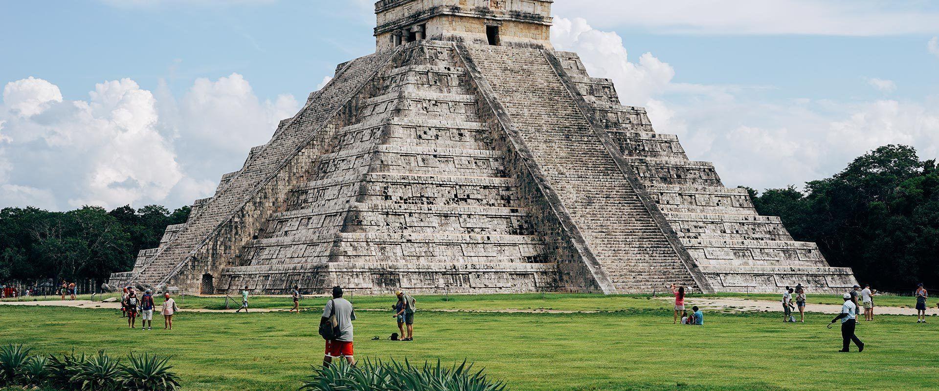 A large stone step pyramid stands on a green lawn with tourists scattered around its base under a partly cloudy sky.