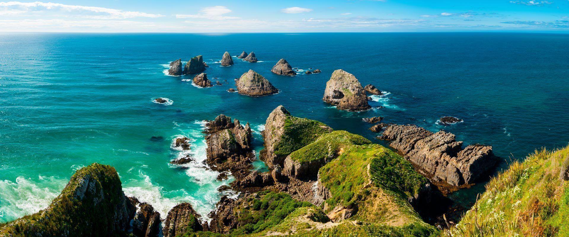 A view from a clifftop shows large rock formations rising from a turquoise and deep blue sea along a rugged coastline.