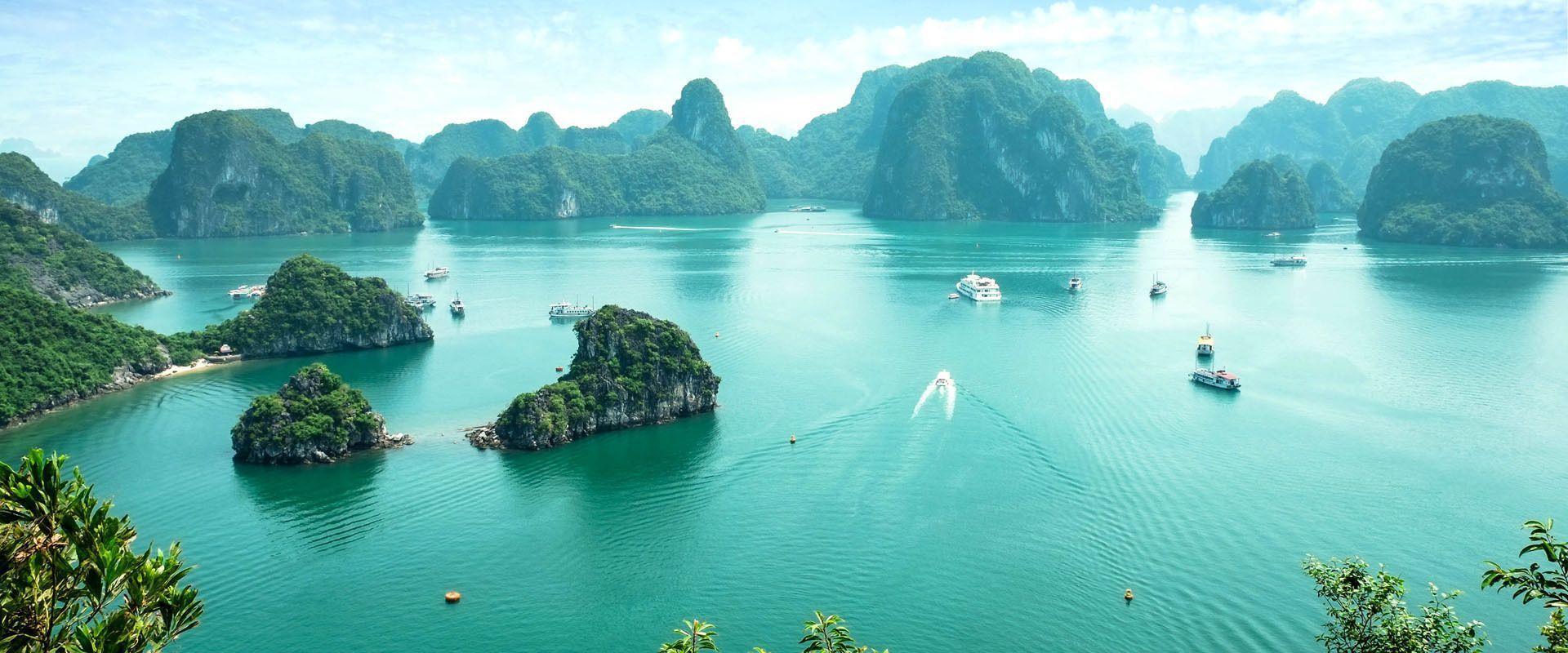 A high-angle view of boats sailing on turquoise water among large, lush green limestone islands under a blue sky.