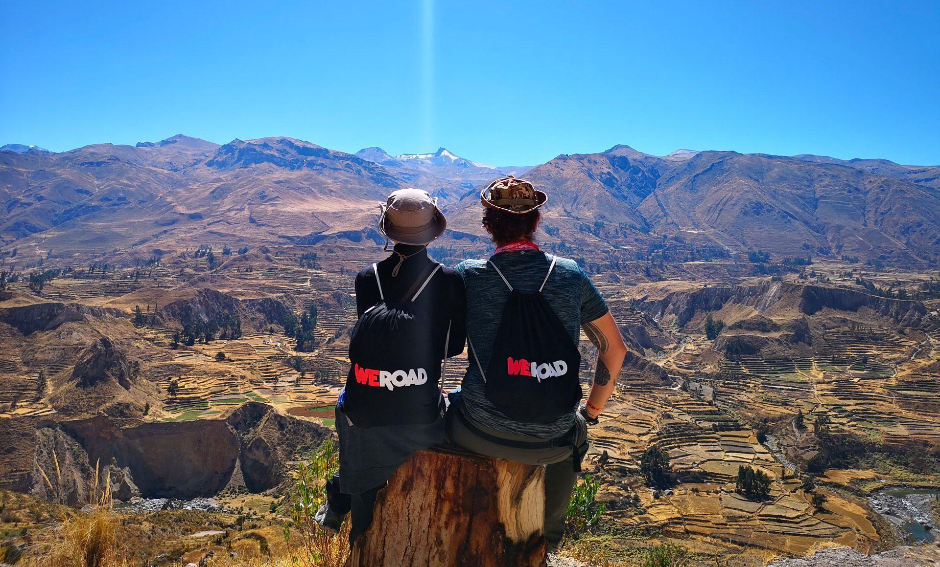 Two people from a WeRoad group trip sitting on a tree stump, overlooking a vast mountain valley with terraced fields.