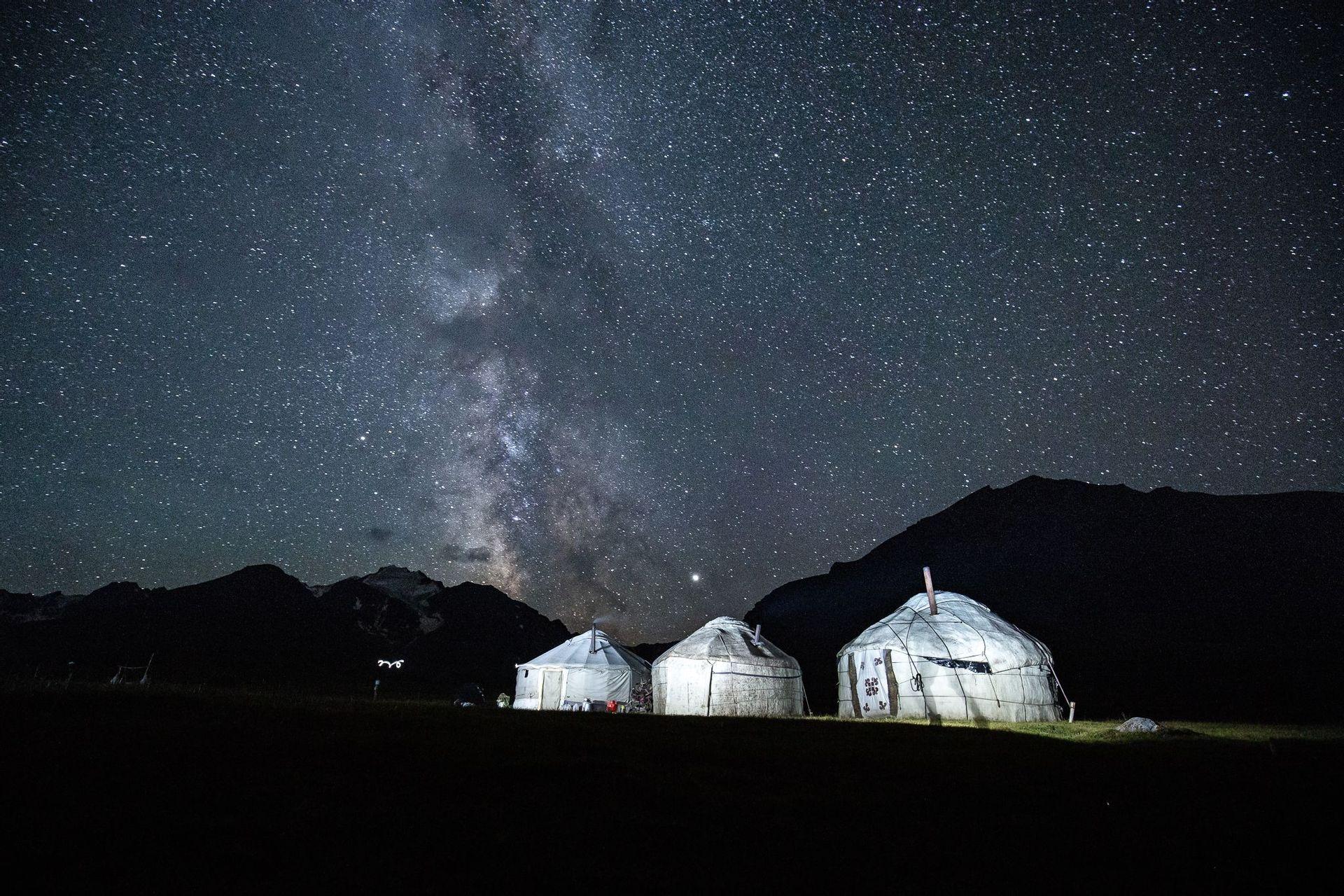 Tre yurte illuminate riposano in un campo ai piedi delle montagne sotto un cielo stellato con la Via Lattea.