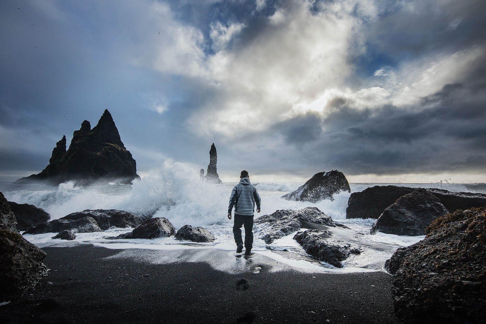 Une personne vue de dos marche sur une plage de sable noir vers une mer orageuse avec des vagues s'écrasant contre des formations rocheuses.