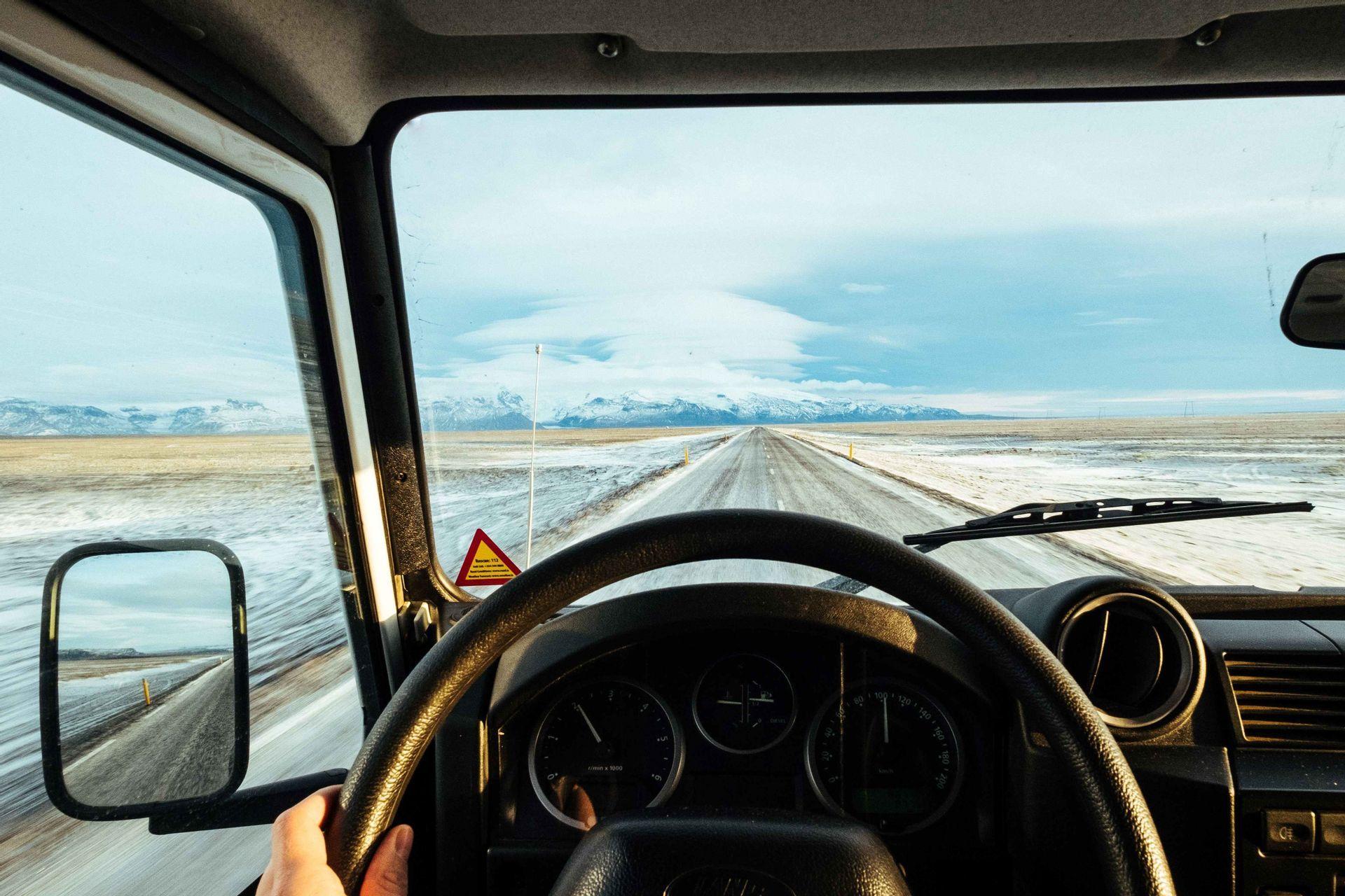 Blick aus dem Auto auf eine gerade, eisige Straße, die durch eine flache, verschneite Landschaft in Richtung ferner Berge führt.