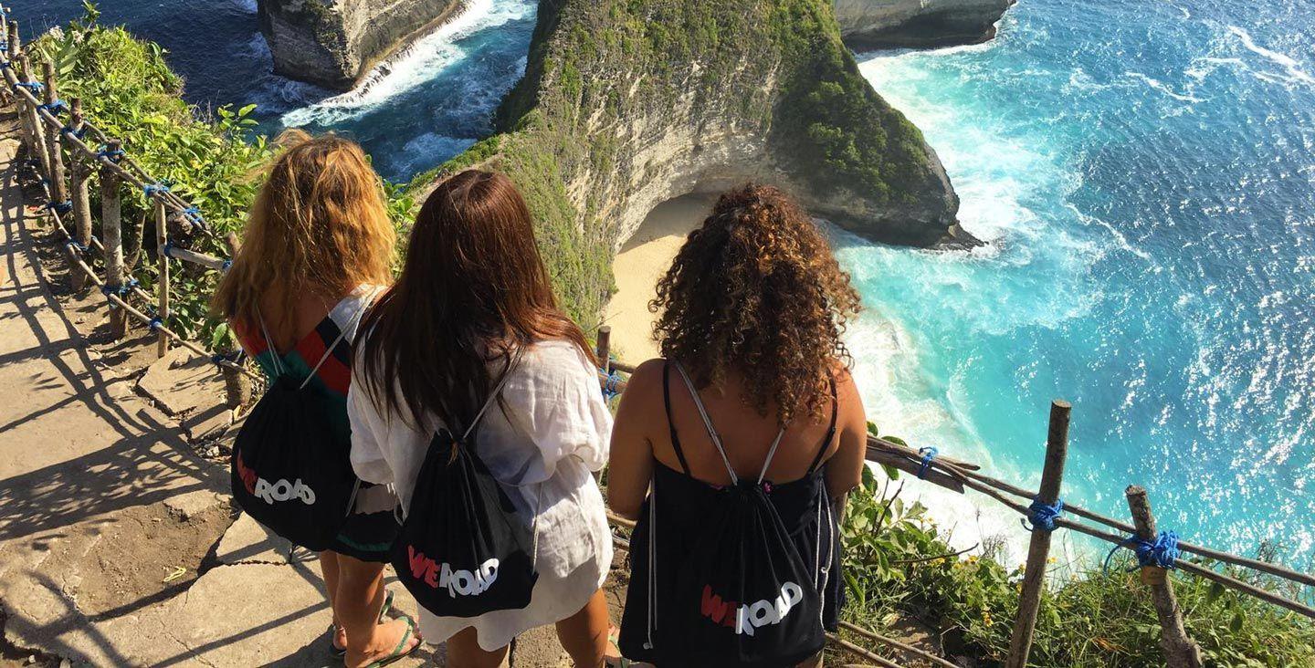 Trois femmes d'un voyage en groupe WeRoad vues de dos, debout sur une falaise surplombant une plage isolée aux eaux turquoises.