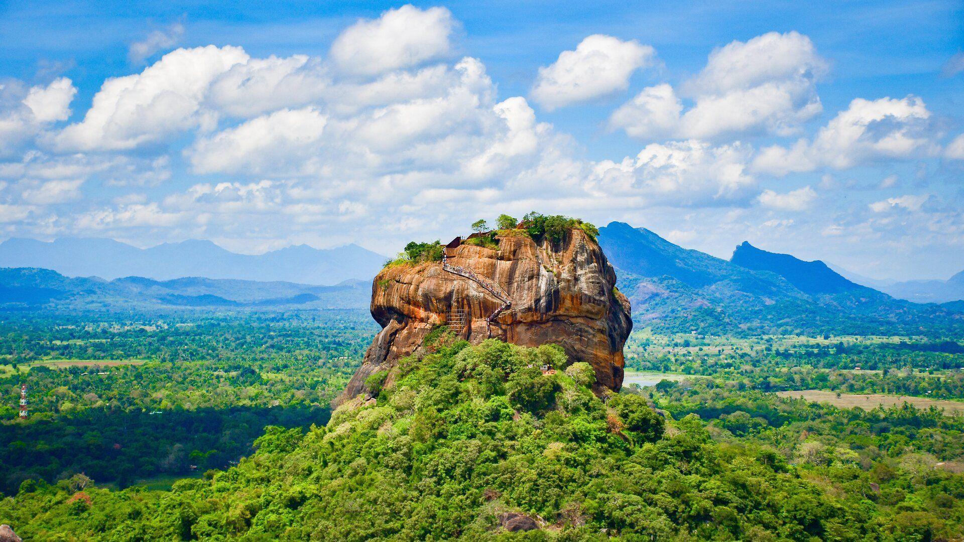 A large rock formation with a visible staircase on its side rises above a vast green jungle under a blue sky with clouds.