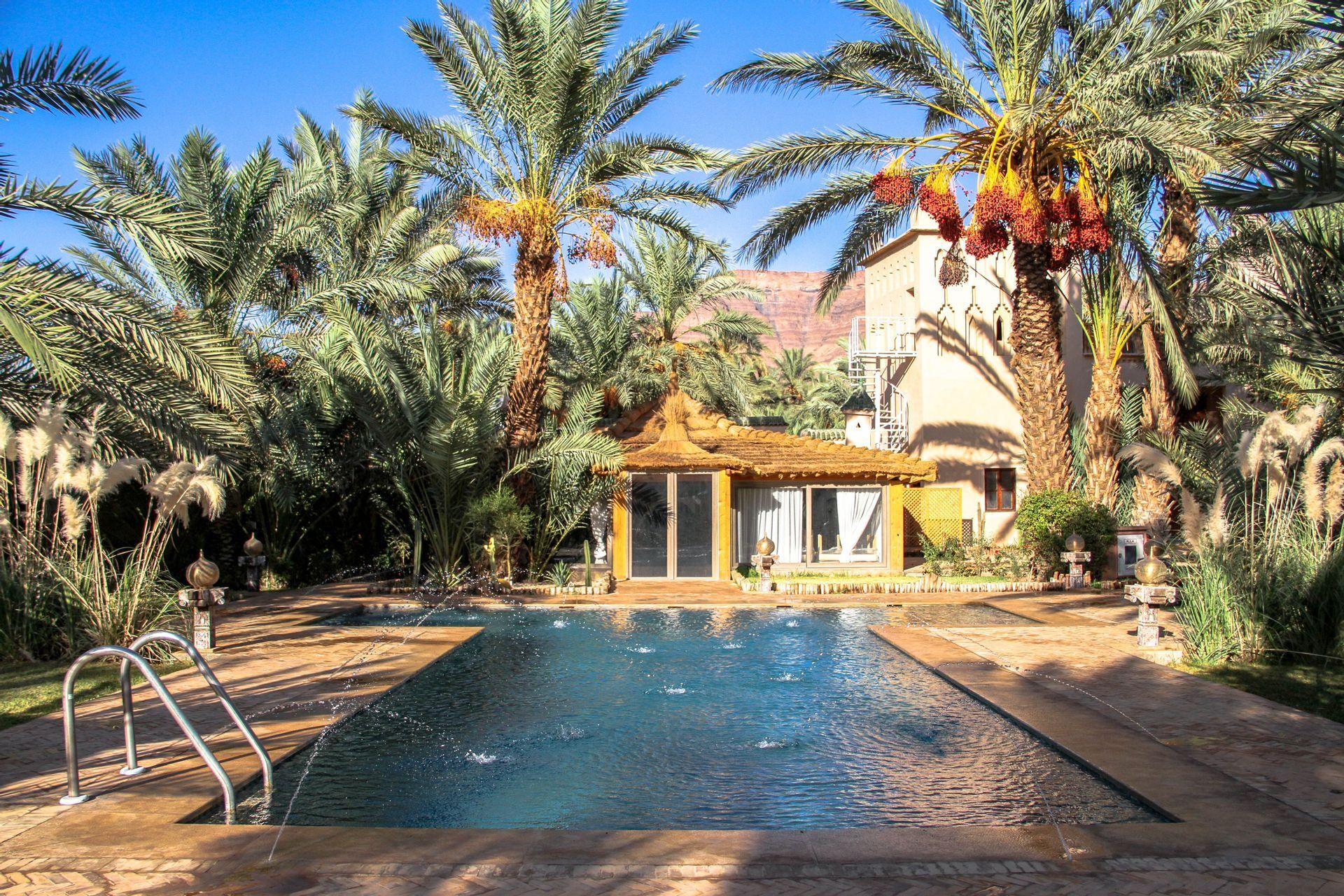 Ein Swimmingpool mit kleinen Springbrunnen liegt in einem sonnigen Innenhof, umgeben von üppigen Palmen und Gebäuden.
