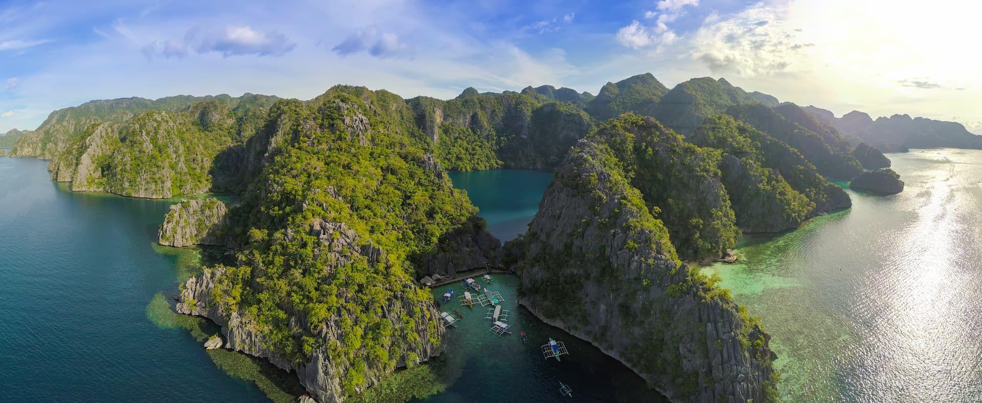 Panoramica aerea di un arcipelago tropicale con isole rocciose verdeggianti, acque turchesi e limpide e diverse barche in una baia.