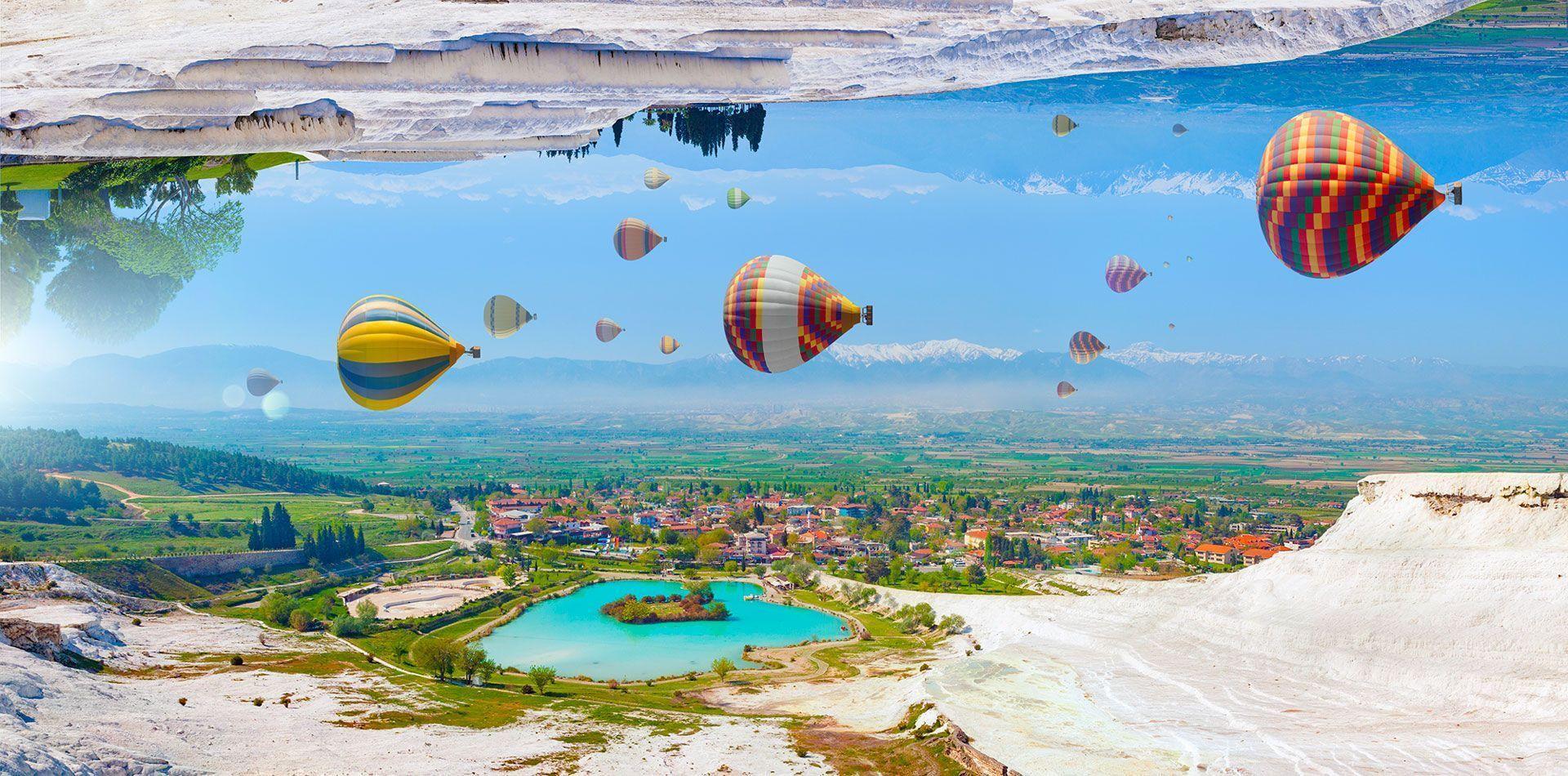 Un paesaggio surreale e capovolto mostra mongolfiere colorate fluttuare sopra una città e un lago turchese, con terrazze bianche riflesse in alto.