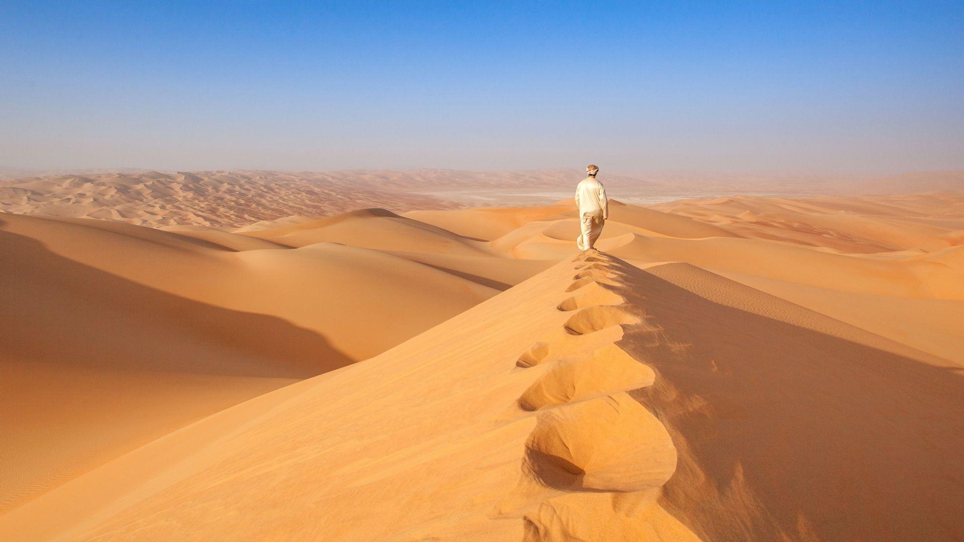 Una persona in abiti tradizionali cammina lungo la cresta di una grande duna di sabbia nel deserto, lasciandosi dietro delle impronte.