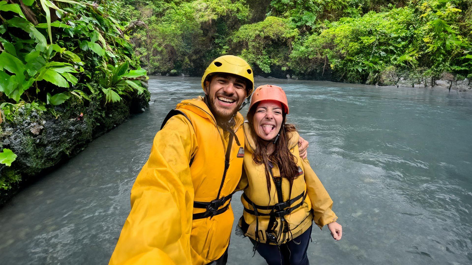 Un uomo e una donna di un viaggio di gruppo WeRoad sorridono per un selfie indossando giubbotti di salvataggio gialli e caschi su un fiume.