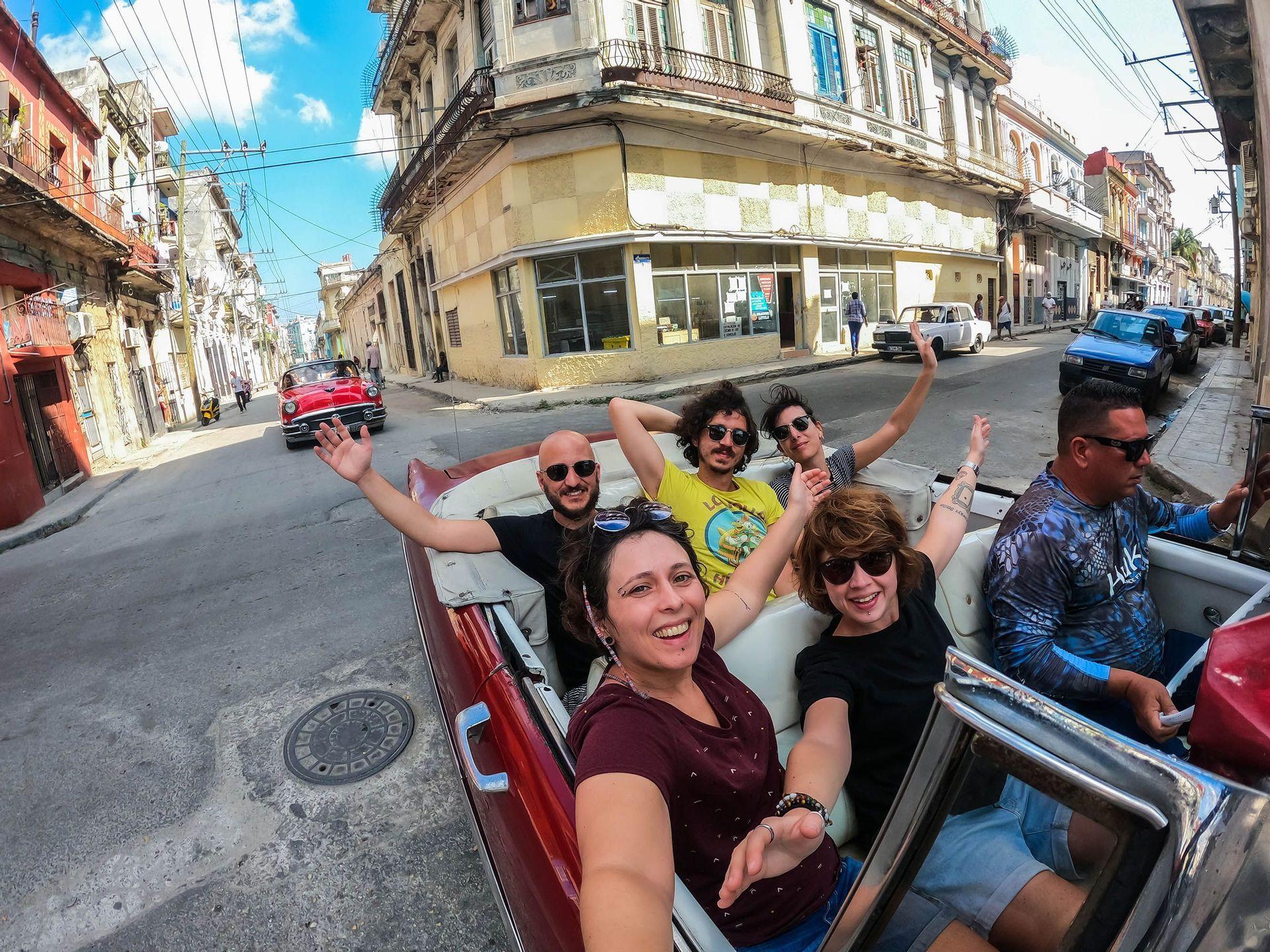 Un grupo de WeRoad se toma una selfie sonriente en un descapotable rojo vintage, circulando por una calle de la ciudad.