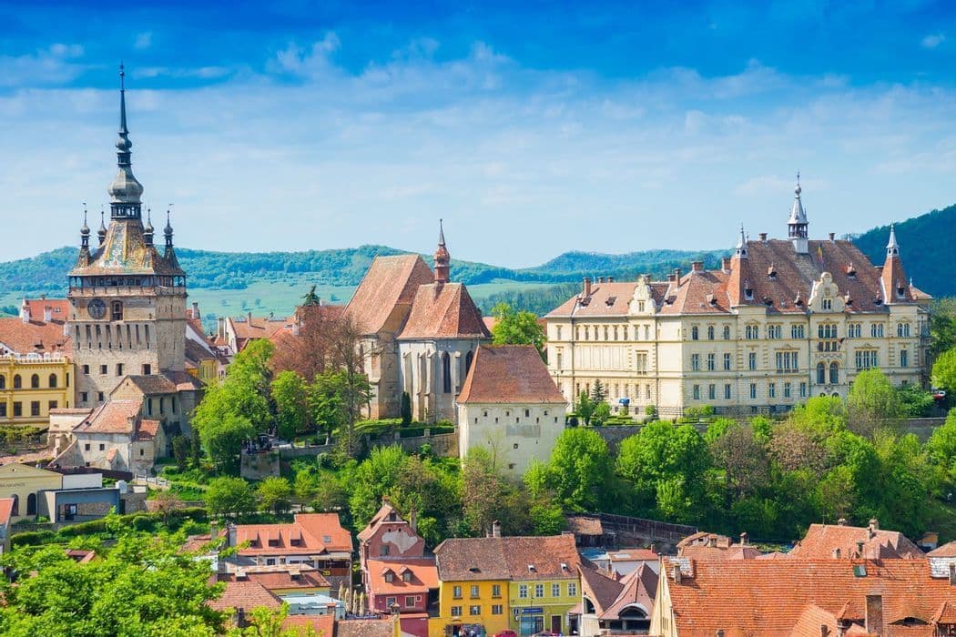viajes-en-grupo-rumania-transilvania-dracula Un pueblo histórico con una torre del reloj prominente y edificios de tejados rojos, enclavado entre verdes colinas bajo un cielo azul.