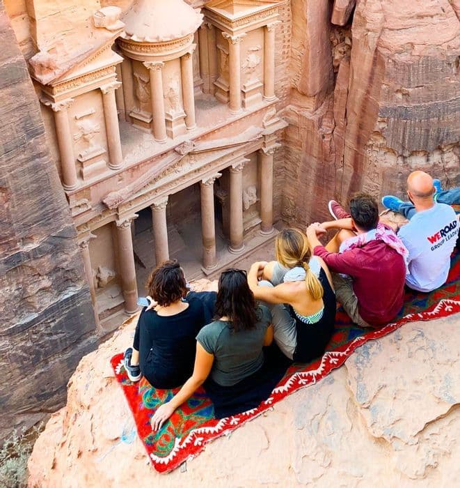 Viaggi over 35 Un gruppo WeRoad è seduto su un tappeto colorato in cima a una scogliera, osservando una facciata antica scolpita nella roccia.