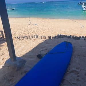 Una tavola da surf blu giace su una spiaggia sabbiosa, con vista sull'oceano turchese e barche sotto un cielo limpido.