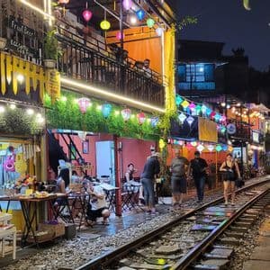 Gente caminando y sentada en cafeterías a lo largo de una vía de tren que atraviesa una calle estrecha e iluminada con faroles por la noche.