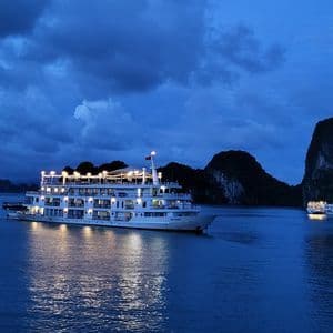 Un crucero iluminado navega en aguas tranquilas al anochecer, rodeado de islas rocosas y oscuras bajo un cielo nublado.