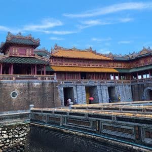 Un gran y ornamentado palacio asiático con techos de tejas amarillas y verdes se alza detrás de una muralla fortificada y un foso bajo un cielo azul claro.