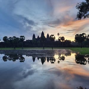 Una silueta de templo y palmeras se reflejan en un lago al amanecer, bajo un cielo con nubes azules, grises y naranjas.