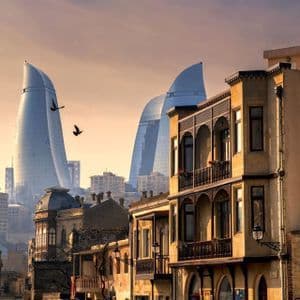 Uno skyline cittadino giustappone edifici tradizionali in pietra con moderni grattacieli di vetro curvi sotto un cielo sfumato al tramonto.