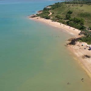 Vista aerea di una spiaggia sabbiosa lungo una costa verde e collinare, con persone che nuotano in acque calme e turchesi.