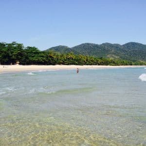 Acqua limpida dell'oceano lambisce una spiaggia di sabbia bianca, incorniciata da alberi verdi rigogliosi e montagne sotto un cielo blu.