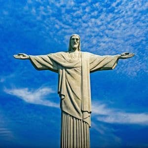 La statua del Cristo Redentore si erge con le braccia aperte contro un cielo blu vibrante punteggiato di nuvole bianche.