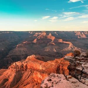 A vast, layered canyon with red rock formations illuminated by golden hour light under a blue sky with clouds.