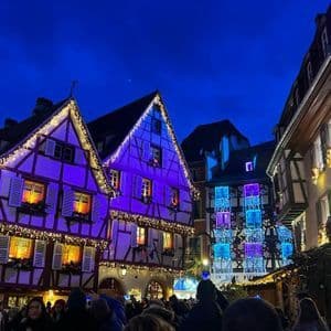 Edificios de entramado de madera iluminados con luces navideñas moradas y blancas sobre una calle concurrida por la noche