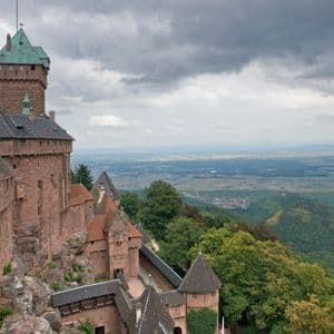 Un castillo de piedra roja con torres y muros fortificados domina un vasto valle verde desde una colina boscosa bajo un cielo nublado.