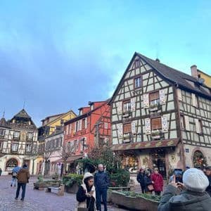 Un grupo de personas camina por una plaza adoquinada rodeada de coloridos edificios con entramado de madera y decoraciones navideñas.