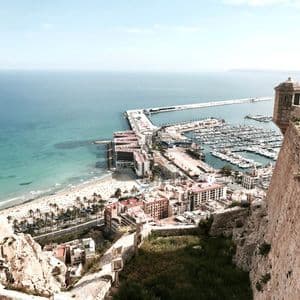 Veduta dall'alto da una fortezza in pietra che domina una città costiera, un porto turistico animato con imbarcazioni e una spiaggia di sabbia.