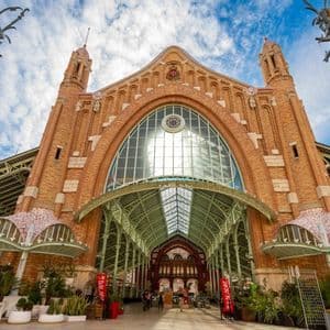 Una veduta dal basso di un elaborato edificio in mattoni con un ampio ingresso ad arco e tetto in vetro, sullo sfondo un cielo blu e nuvoloso.