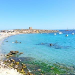 Una baia costiera soleggiata con una spiaggia sabbiosa, acqua cristallina turchese, diverse imbarcazioni e una torre di pietra su un promontorio lontano.
