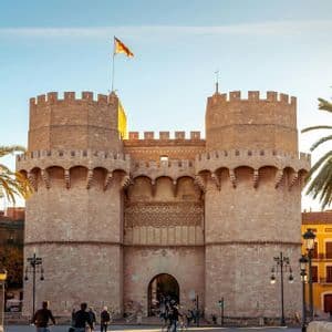 Un grande portale in pietra storico con due torri merlate, mentre la gente passeggia e va in bicicletta nella piazza soleggiata sottostante.