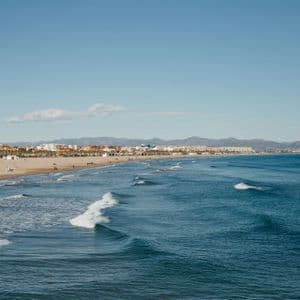 Le onde si infrangono su un'ampia spiaggia sabbiosa delimitata da una città costiera e montagne lontane sotto un cielo azzurro e limpido.
