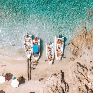 Veduta aerea di un viaggio di gruppo WeRoad con barche ormeggiate su una spiaggia di sabbia, caratterizzata da acque turchesi e cristalline e coste rocciose.