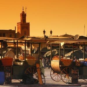 Dos carros de madera llenos de naranjas se encuentran en una bulliciosa plaza de mercado bajo un cielo anaranjado, con un minarete al fondo.