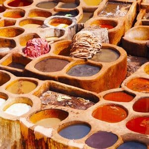 In einer Freiluftgerberei geht ein Mann in einer blauen Djellaba an großen, steinernen Bottichen vorbei, die mit bunten Farbstoffen gefüllt sind.
