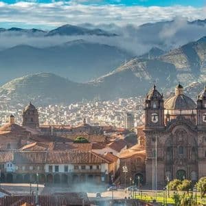 Cattedrali storiche in pietra con cupole e torri dominano una città incastonata in una valle, con montagne avvolte nella nebbia che si stagliano sullo sfondo.