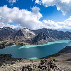 Una vista aerea di un lago alpino turchese adagiato tra una catena montuosa rocciosa con cime innevate sotto un cielo blu e nuvoloso.