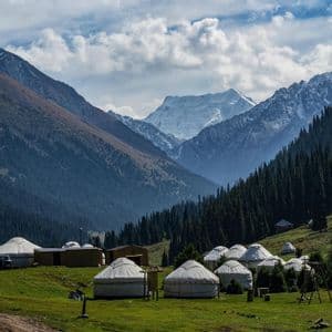 A yurt camp sits on a green hillside, with a backdrop of pine forests and large, snow-capped mountains under a cloudy sky.