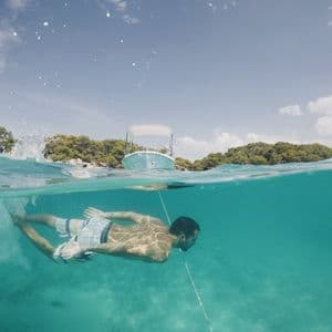 Vue aquatique et aérienne d'un homme nageant sous l'eau dans une mer turquoise et limpide, avec des bateaux et une côte arborée visibles en surface.
