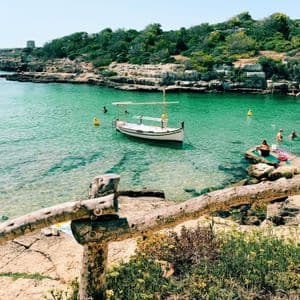 Menschen schwimmen und entspannen in einer ruhigen, türkisfarbenen Bucht mit mehreren kleinen Booten nahe einer felsigen, baumbestandenen Küste.
