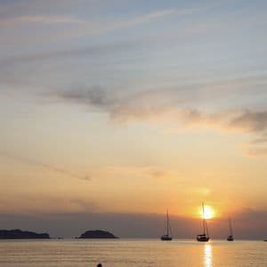 Eine Silhouette im Meer, die einen goldenen Sonnenuntergang mit Segelbooten am Horizont beobachtet.
