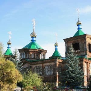 A large wooden Orthodox church with green roofs and golden onion domes surrounded by trees and flowers under a blue sky.