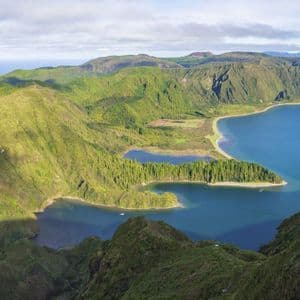 A panoramic view of a deep blue lake inside a volcanic caldera, surrounded by lush green mountains with the ocean in the distance.